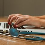 photo of person typing on a laptop computer with a stethoscope on table next to them