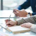 Hands of female making notes in notebook while sitting at desk next to another person working at a computer